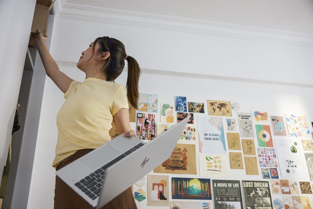 Woman reaching up to a shelf with one hand and laptop computer in other hand